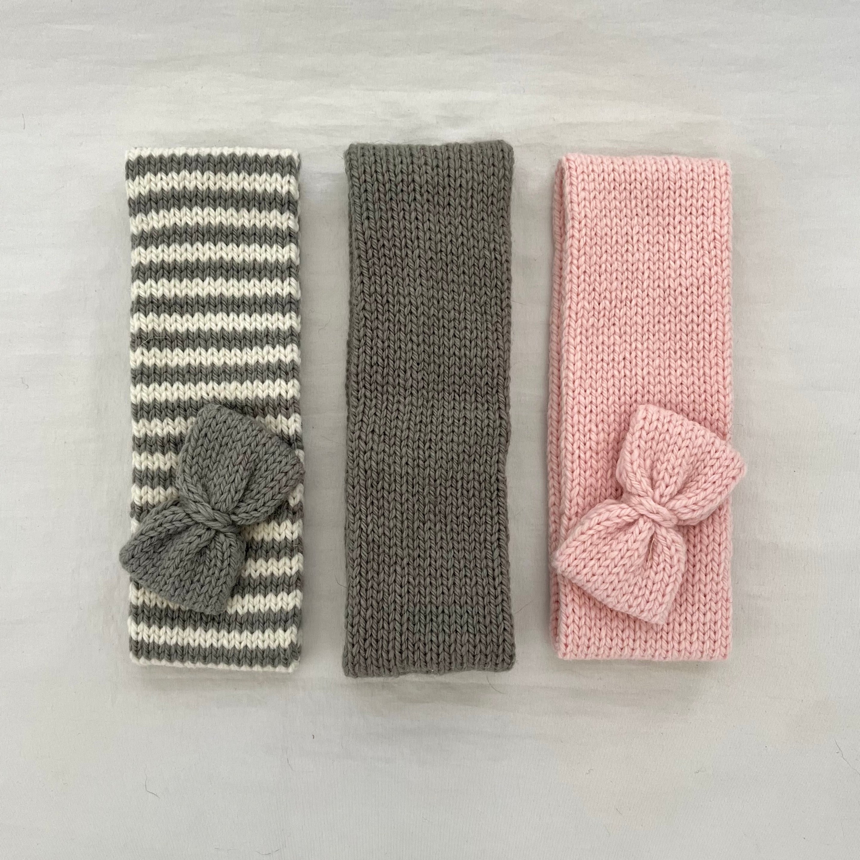 Three knitted baby headbands with bows, laid flat on a light gray surface. The left headband has gray and white stripes with a gray bow, the middle is solid gray, and the right is solid pink with a pink bow.