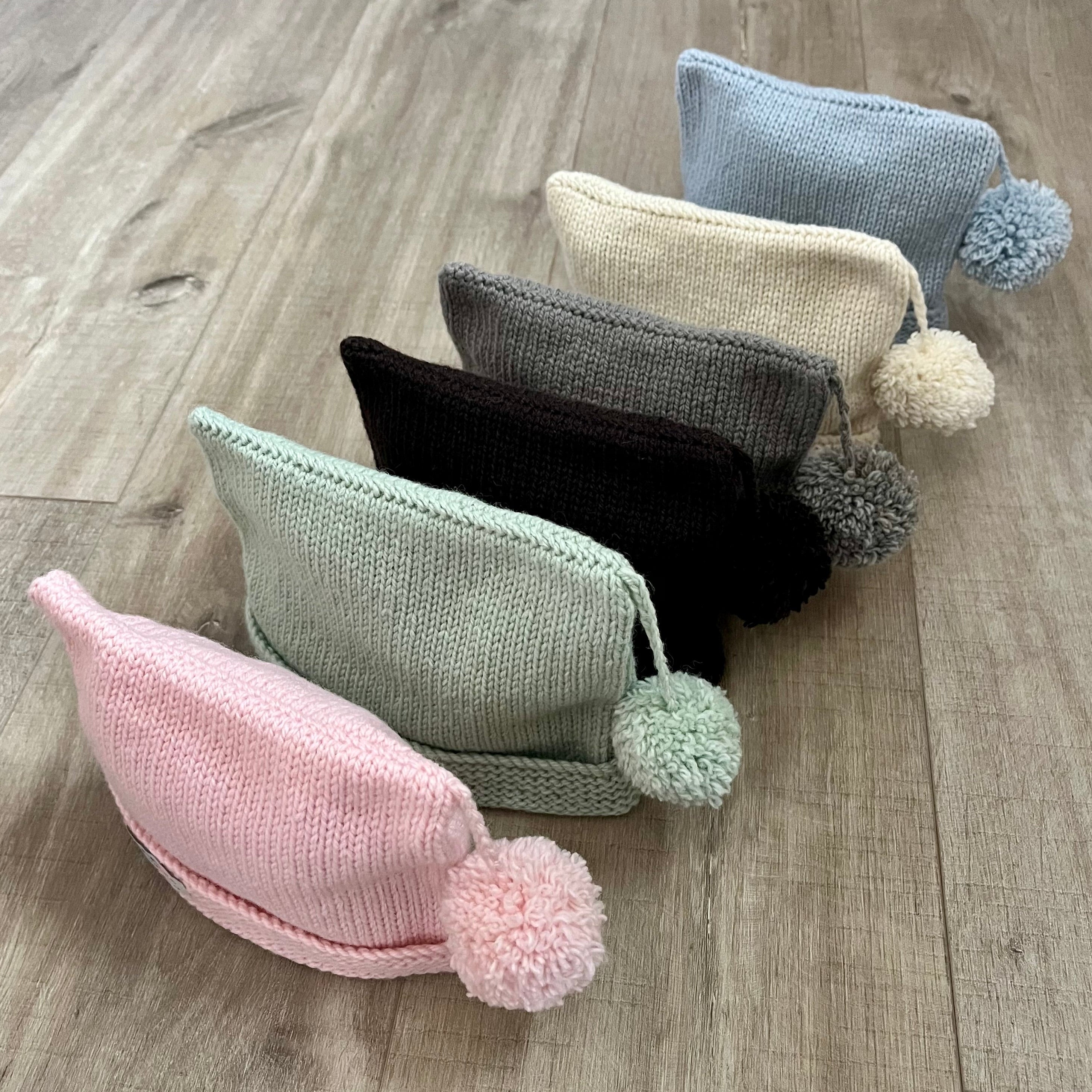 Six knitted baby hats with pom-poms in pink, sage green, brown, charcoal, and light blue, arranged diagonally on a light wood floor.