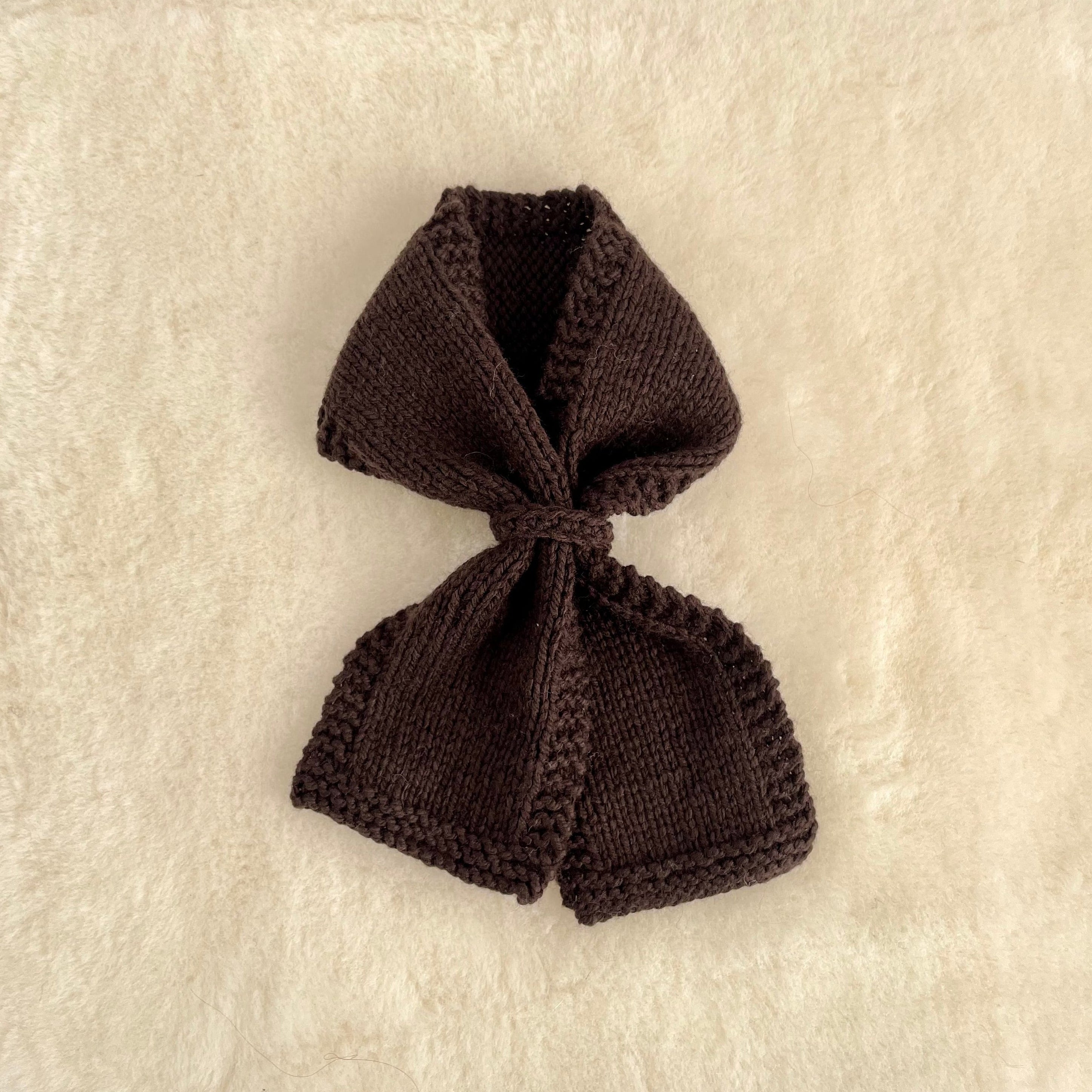 A small knitted loop scarf in brown, laid flat on a textured cream-colored fabric background.