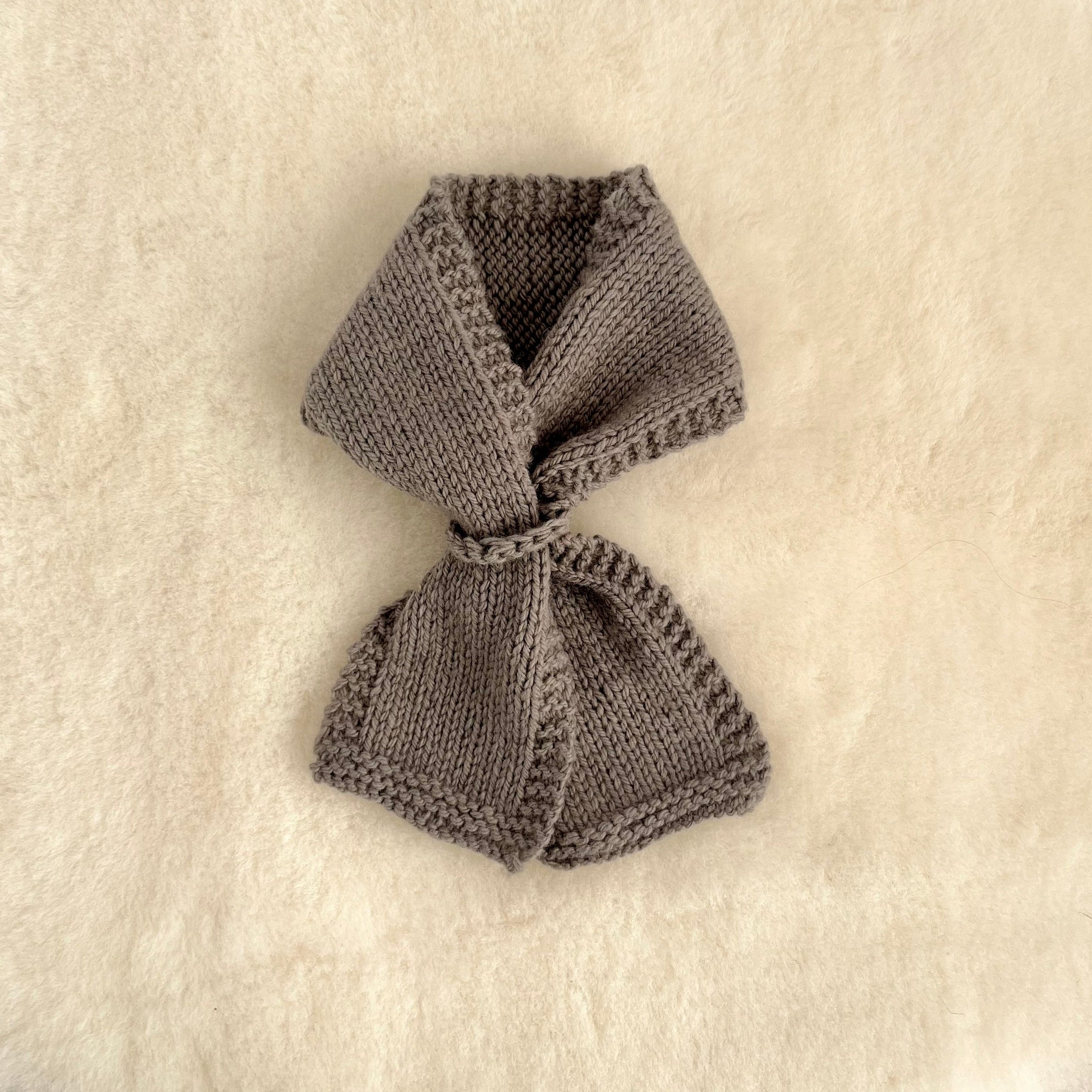A small knitted loop scarf in mushroom, laid flat on a textured cream-colored fabric background.