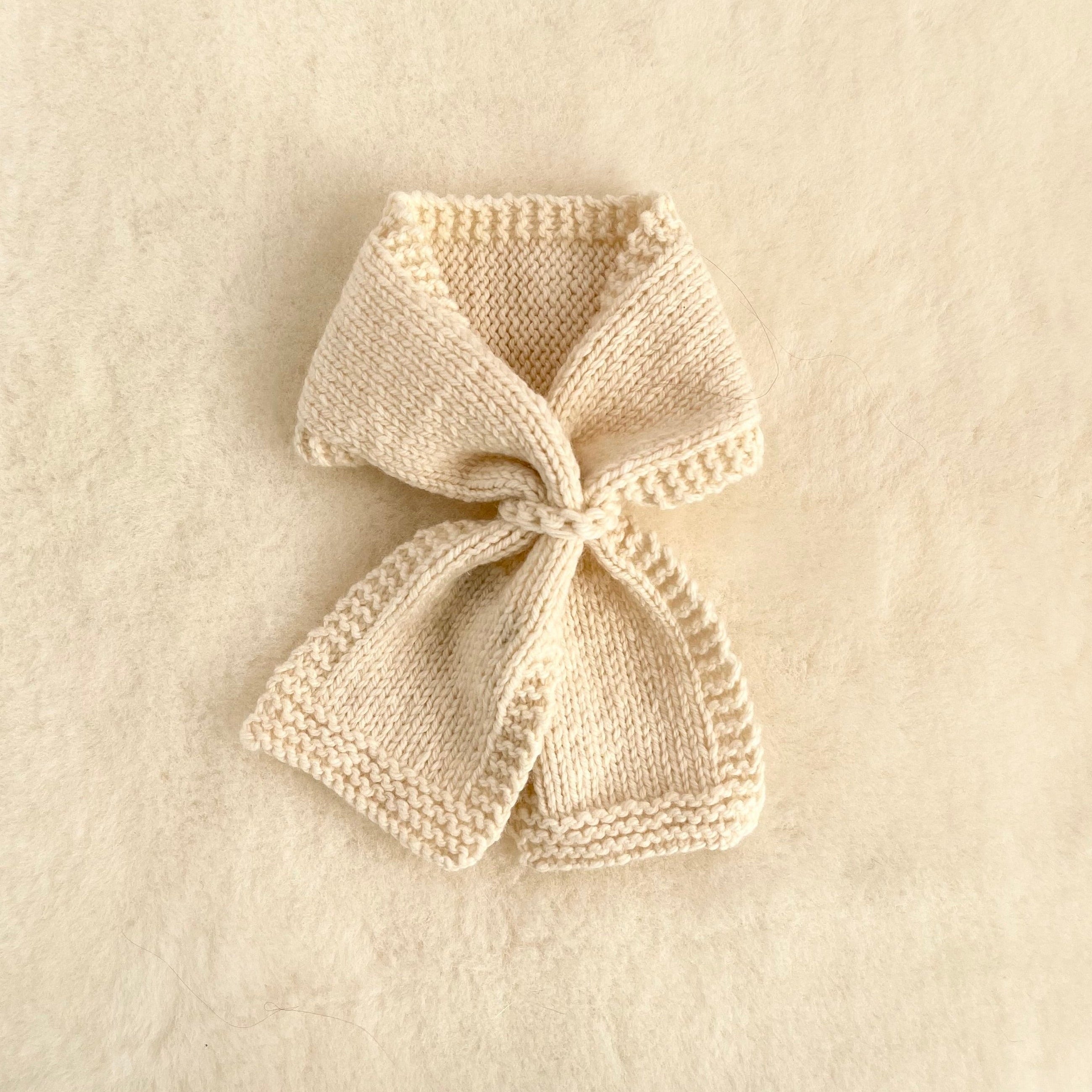 A cream-colored knitted baby scarf with a bow, displayed on a soft, textured cream fabric background.