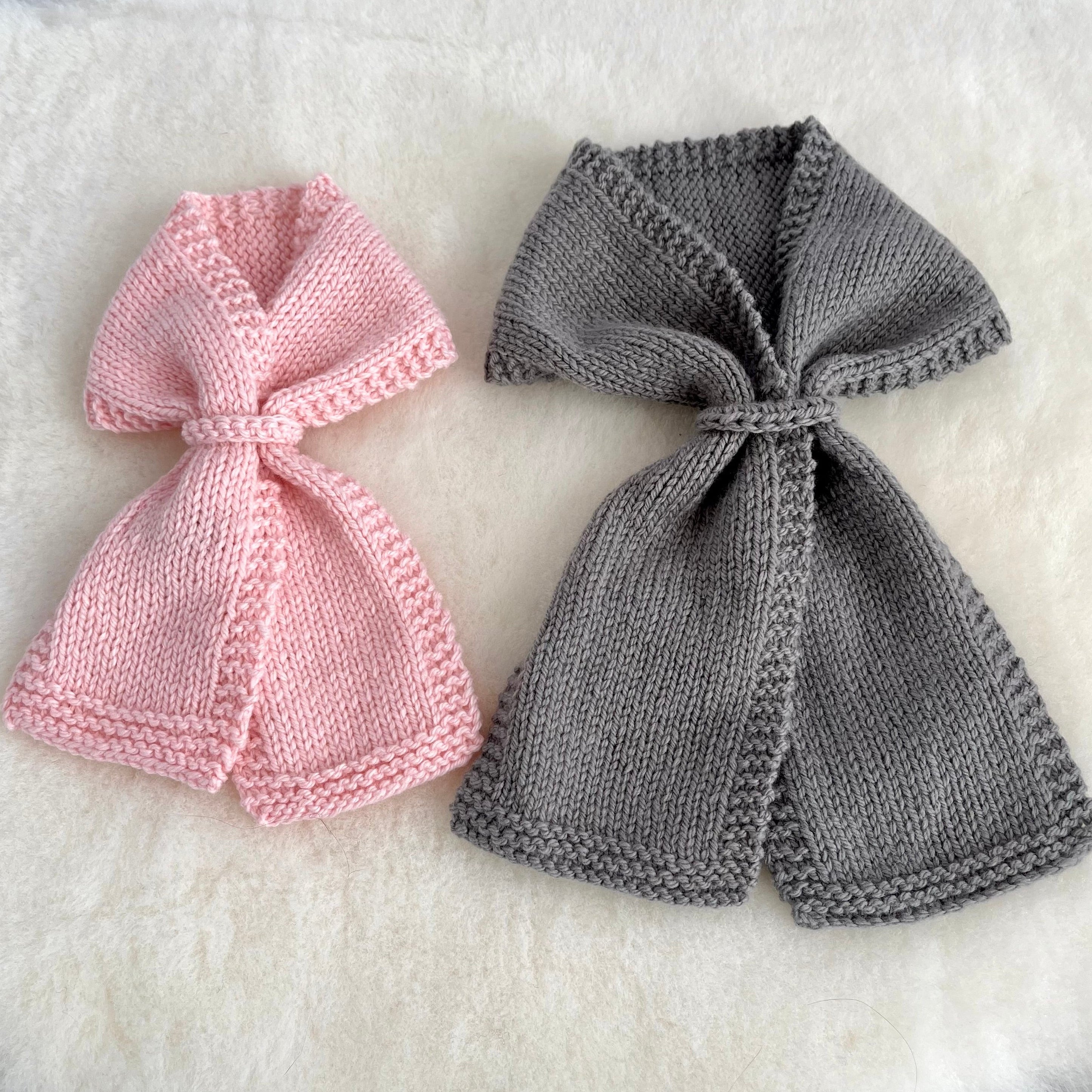 Two knitted bow-shaped scarves, one in light pink and one in medium gray, displayed on a soft white faux-fur surface.