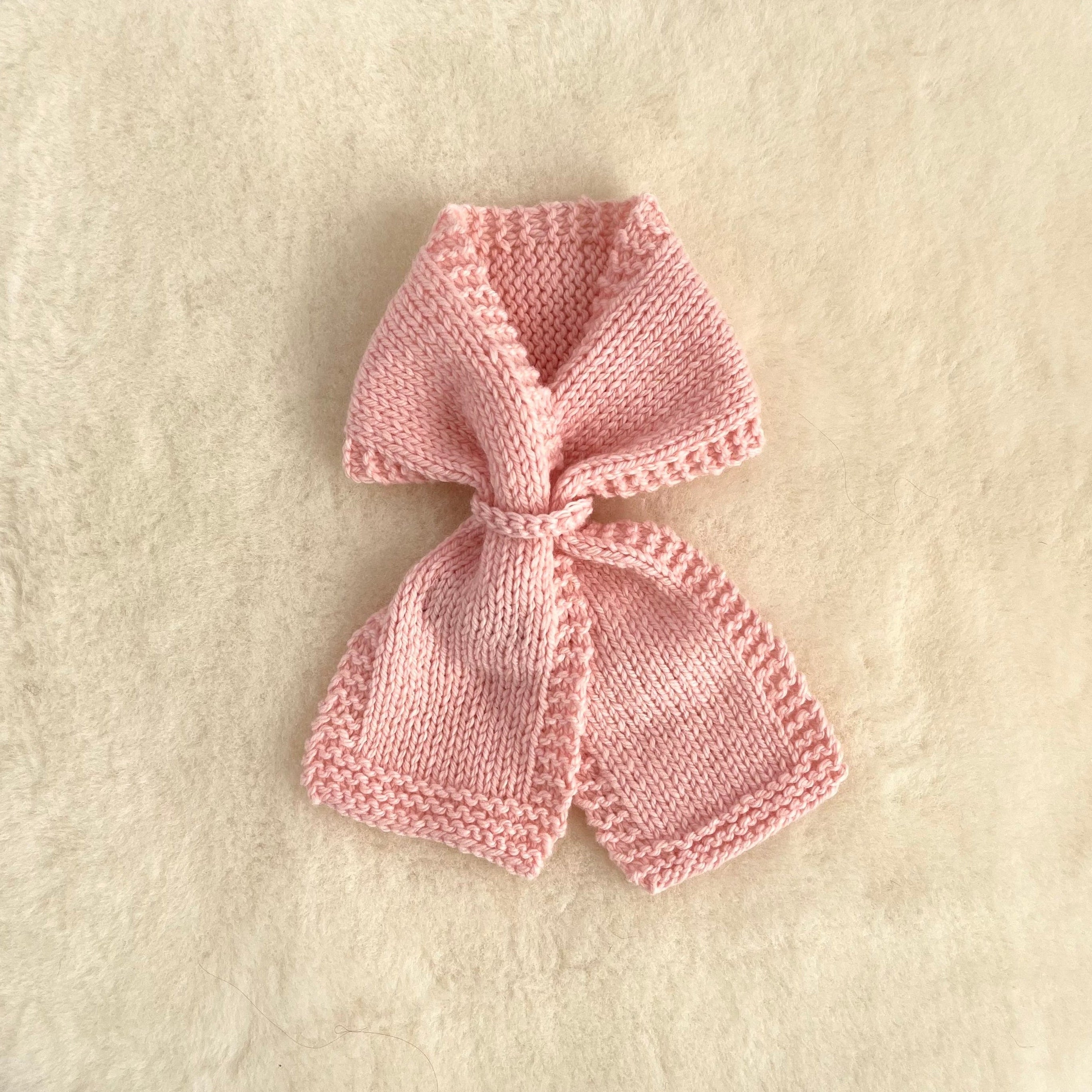 A small knitted loop scarf in soft light pink, laid flat on a textured cream-colored fabric background.