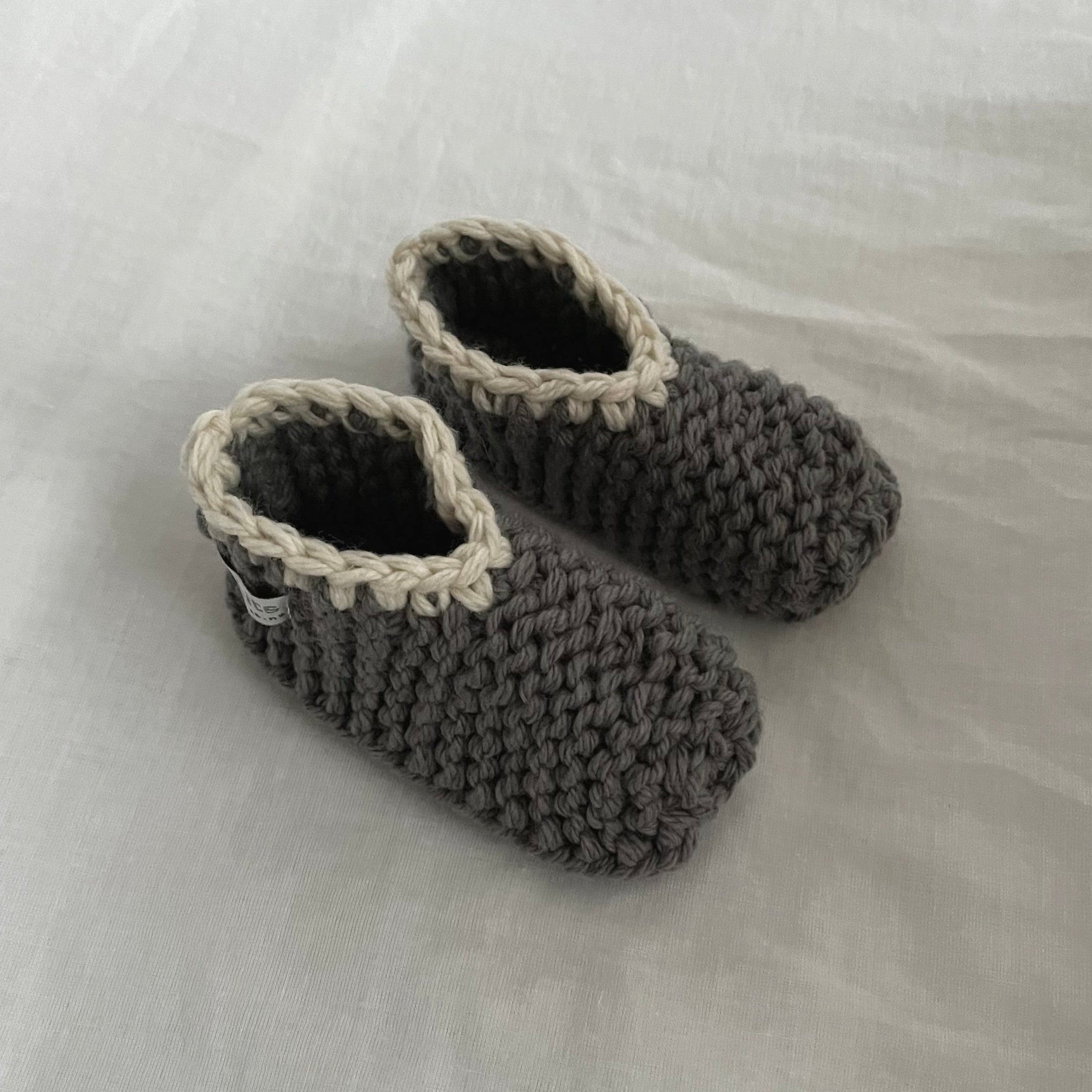 A pair of handmade, mushroom-colored merino slippers with dark gray trim, displayed on a soft, off-white fabric background.