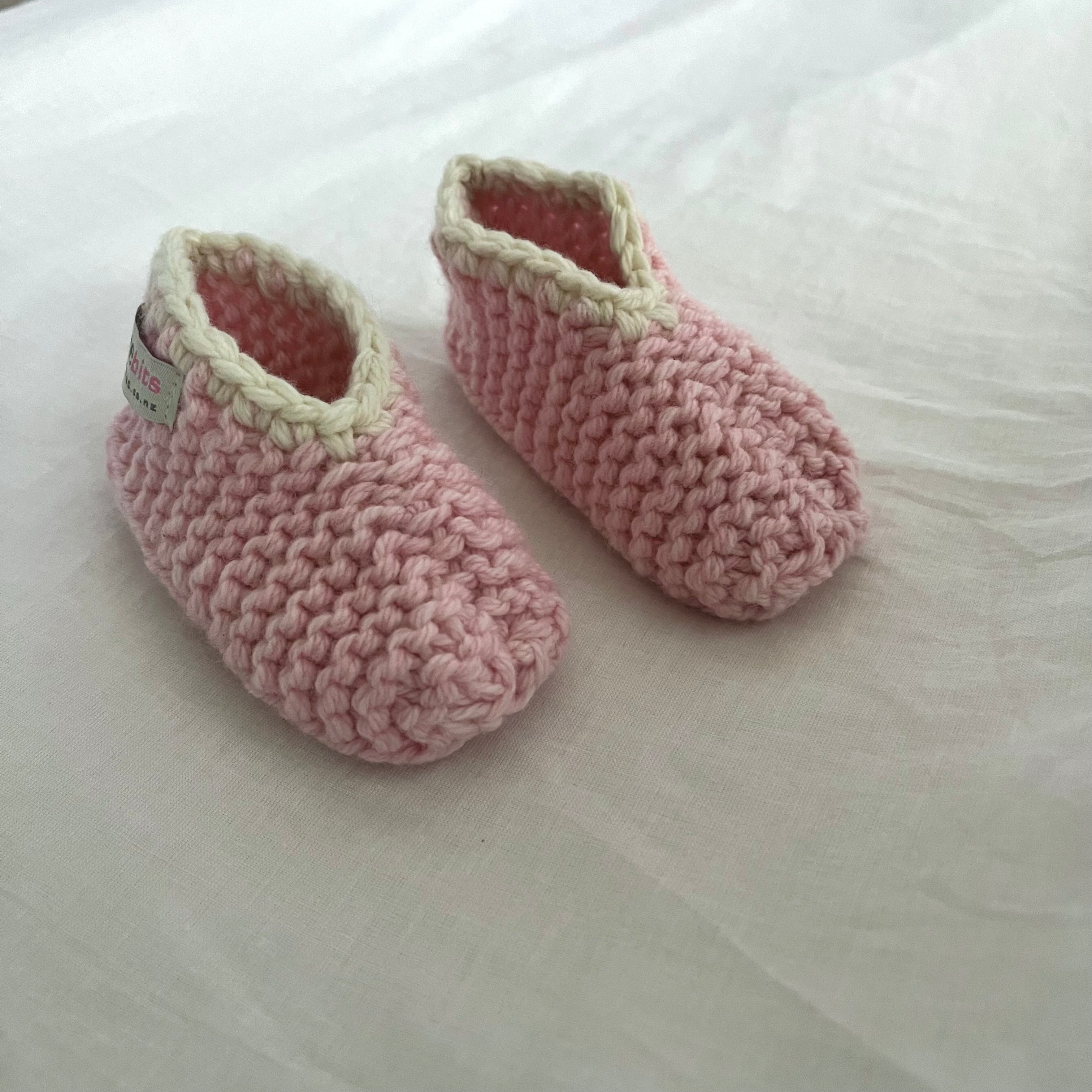 A pair of handmade, pink-colored merino slippers with dark gray trim, displayed on a soft, off-white fabric background.
