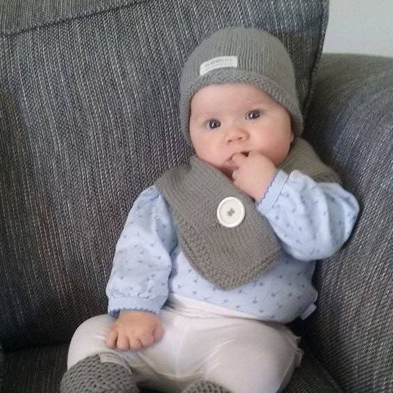 A baby wearing a gray knit beanie and matching gray knit scarf over a light blue patterned long-sleeve top and white pants, sitting on a gray upholstered chair.