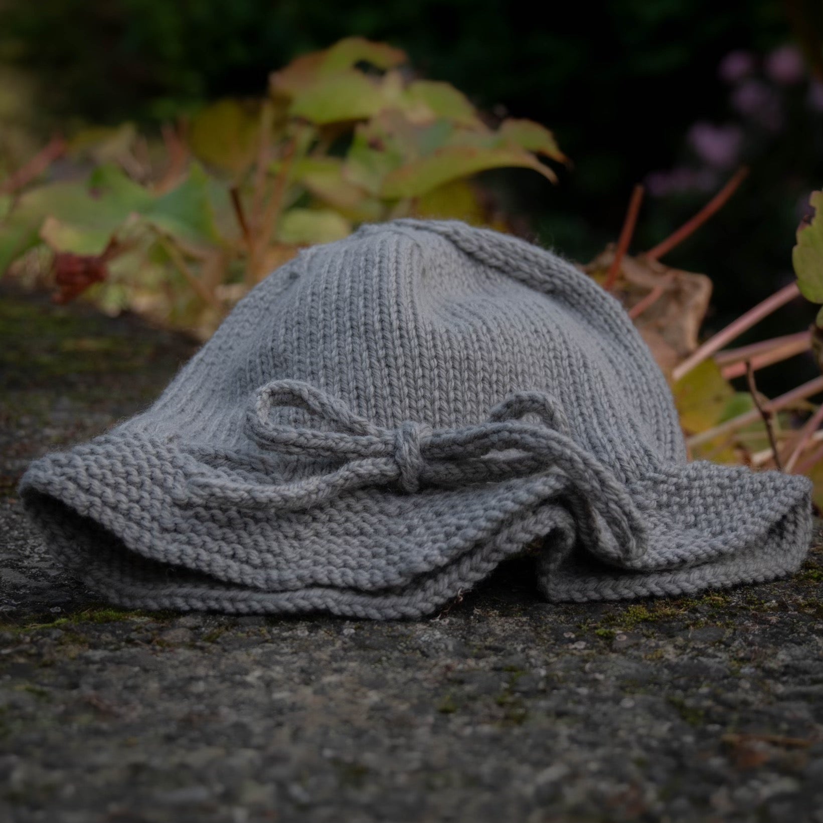 A gray hand-knit sun hat with a bow detail, resting on a textured stone surface with a blurred natural background of green leaves.