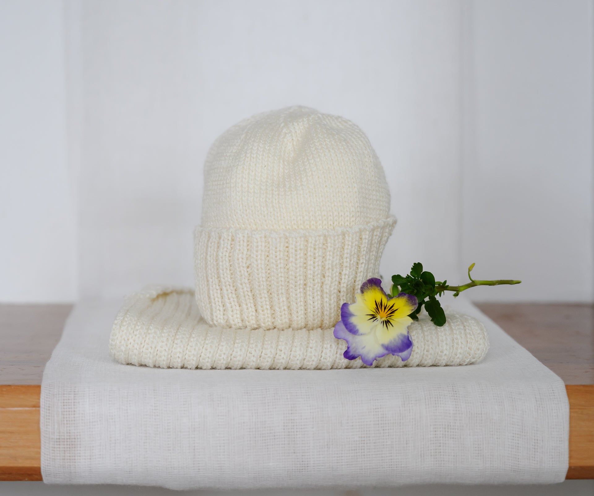 A cream-colored knit beanie hat with a matching wide brim, styled on a white fabric surface with a small purple and yellow pansy flower on the side.