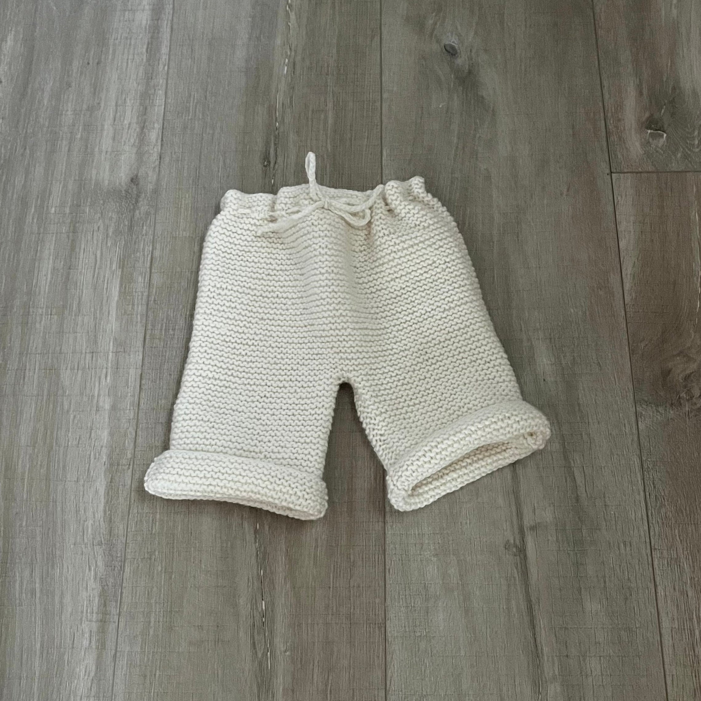 Cream-colored knit baby pants with a drawstring waist and rolled cuffs, laid flat on a gray wood plank floor.