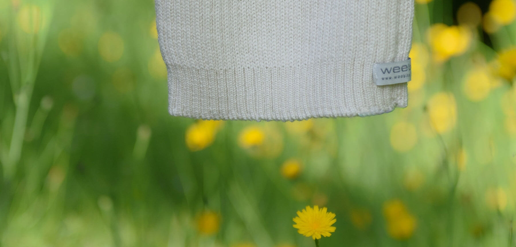 natural knit vest with a brand label hanging in a field of yellow flowers.