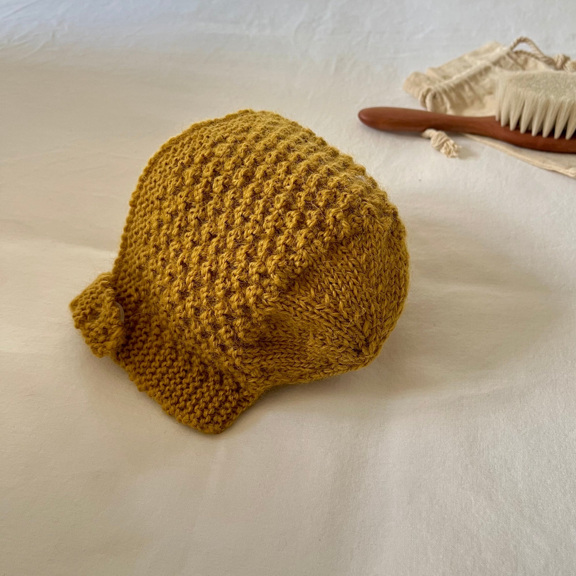 Knitted mustard yellow hat on a white surface with a wooden brush and white bag in the background.