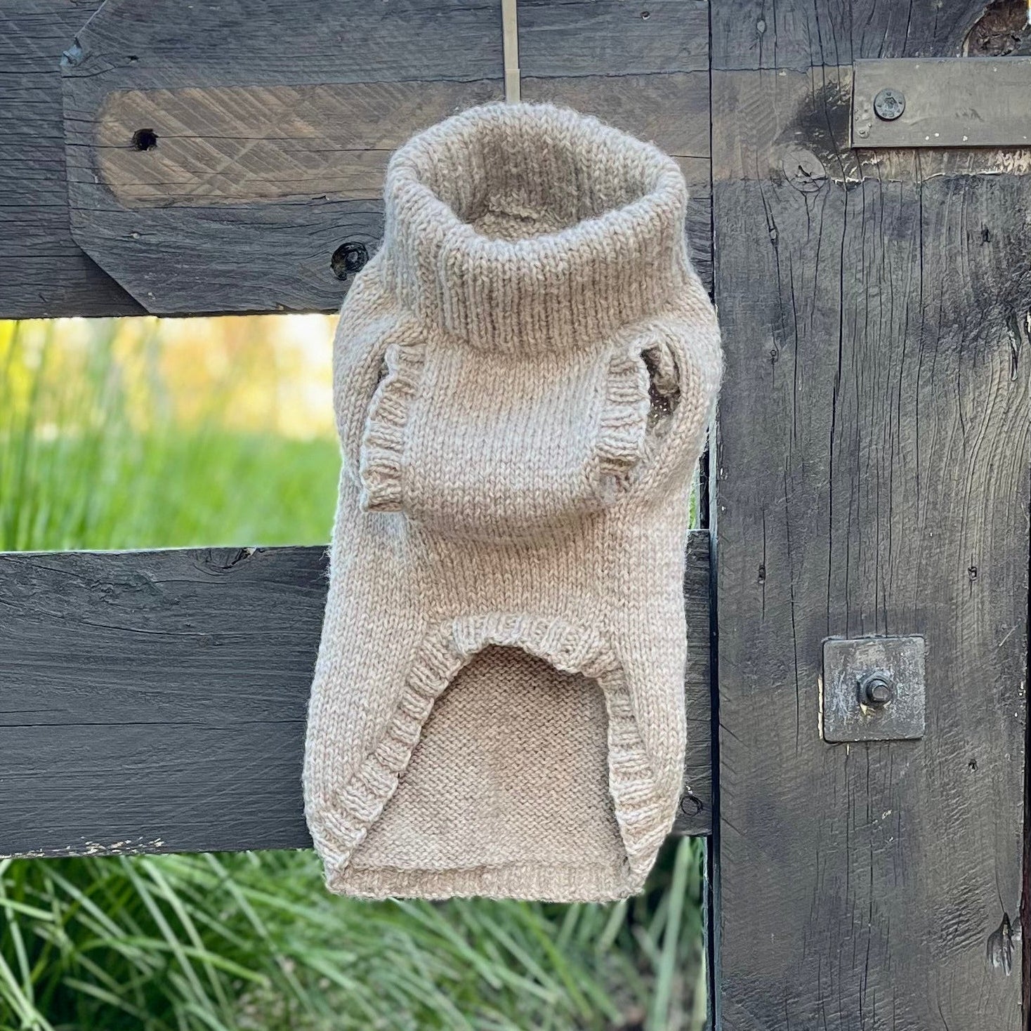 A beige knit pet sweater styled like a turtleneck dog coat, hanging on a rustic dark wooden fence with green grass in the background.