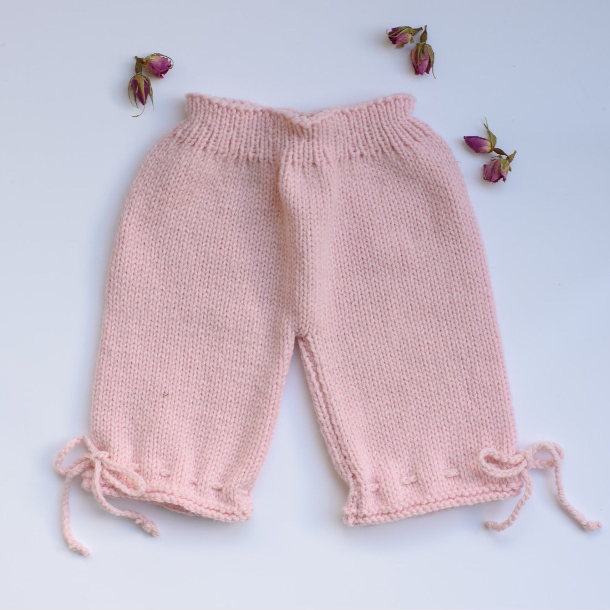 A pair of hand-knit baby pants in a soft light pink color, laid flat on a light blue background with small dried rose buds placed around them.