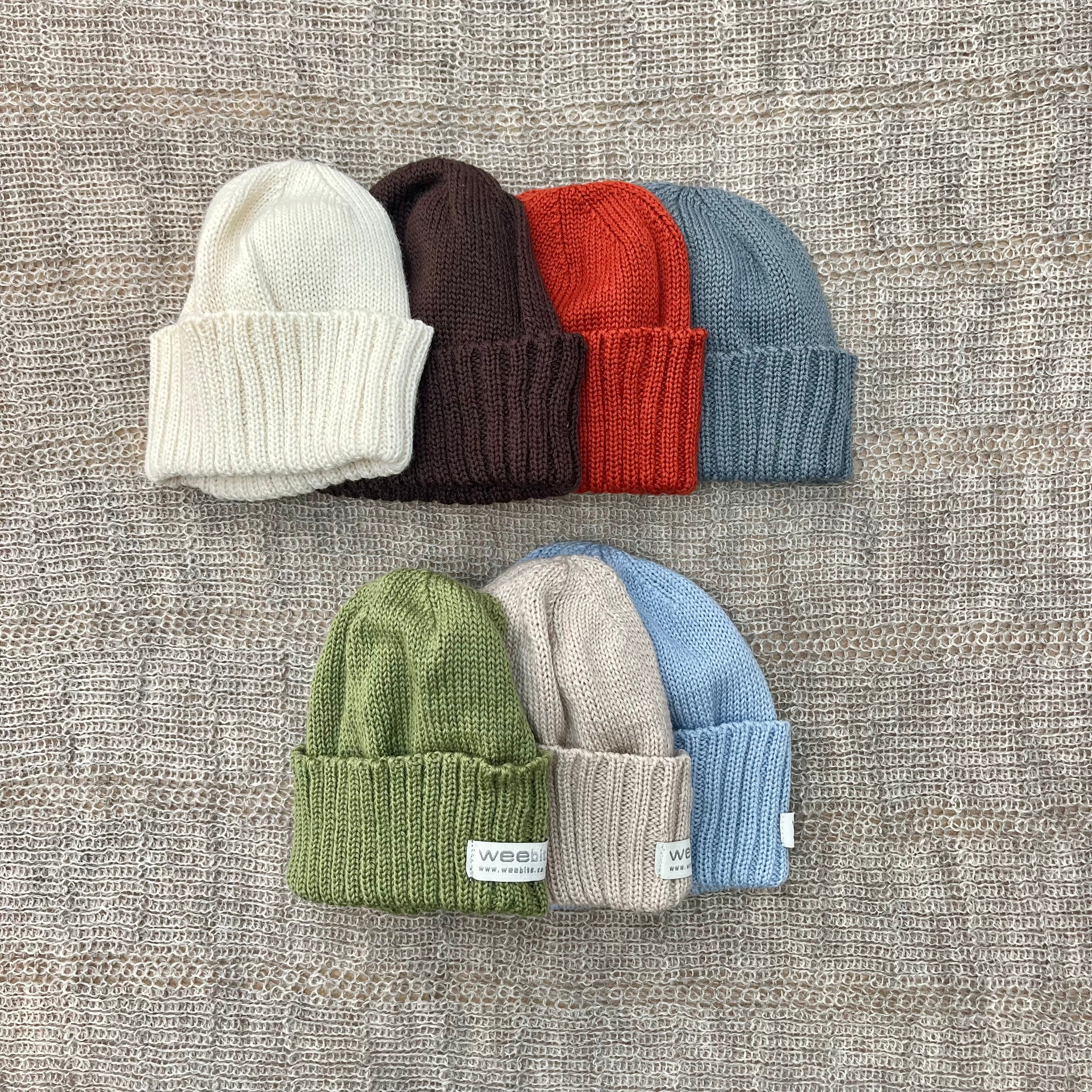 Six ribbed knit beanies arranged in two rows on a textured beige fabric background. The top row shows cream, dark brown, rust red, and slate blue.