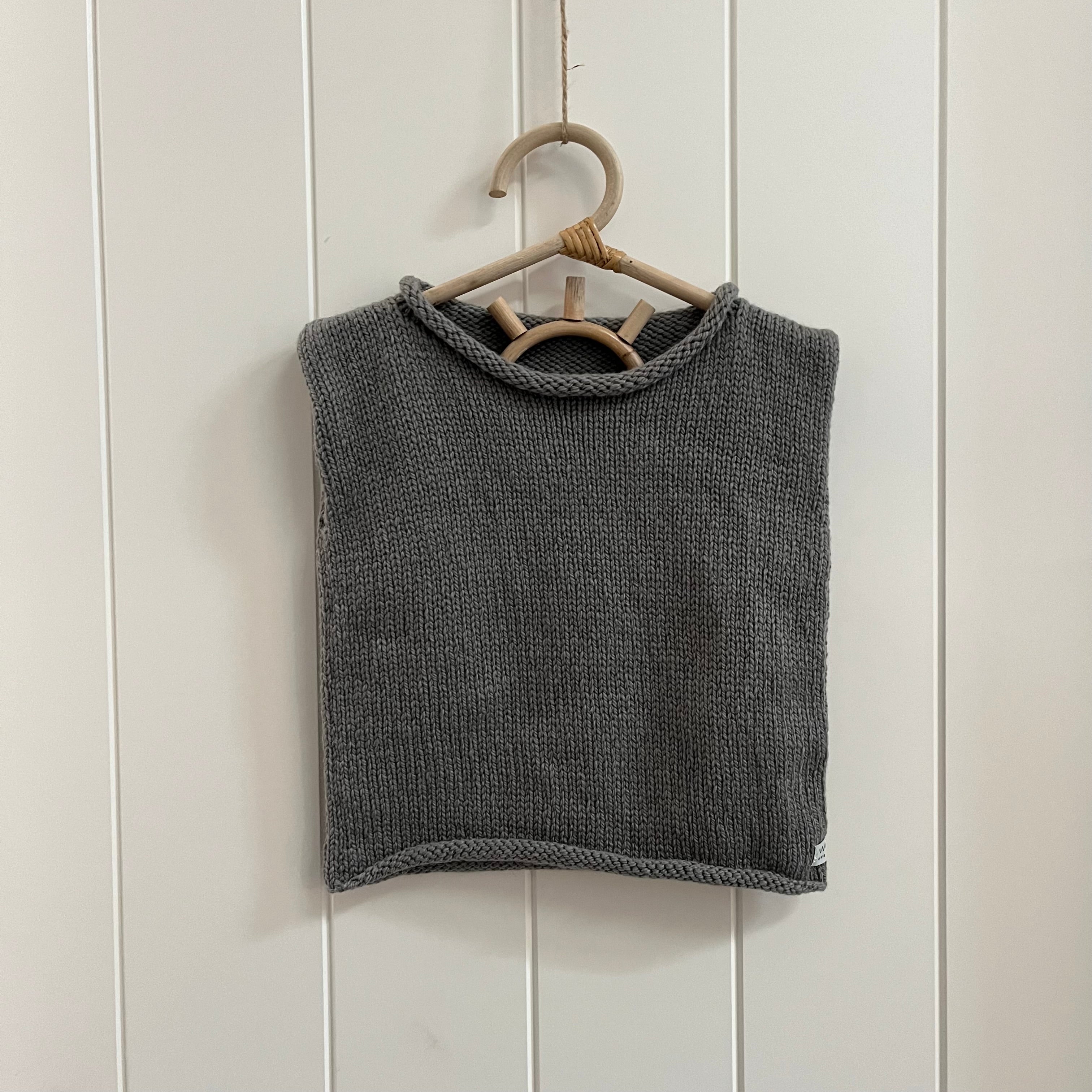 A dark gray knit sleeveless  vest hanging on a light wood hanger against a white paneled wall.