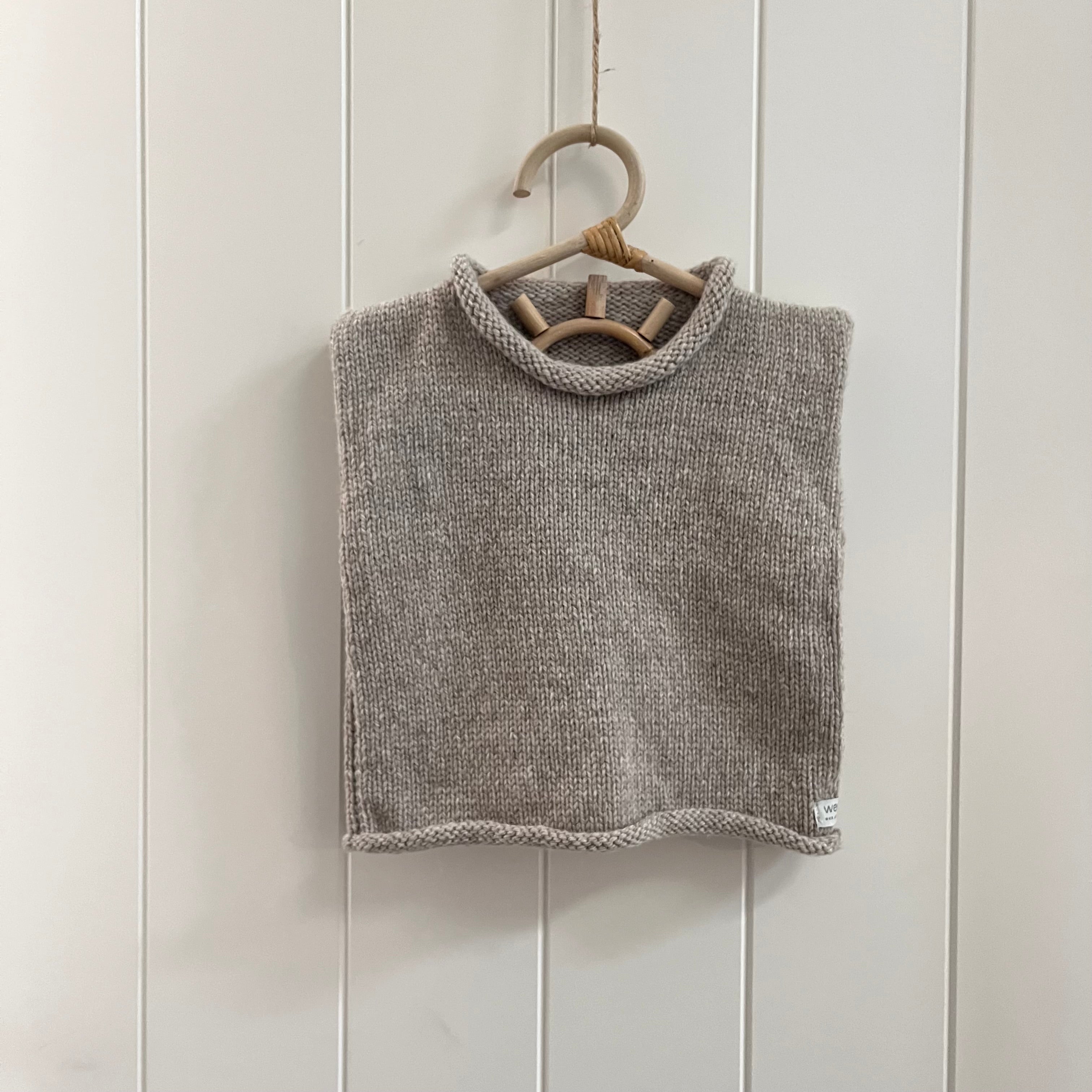 A oatmeal knit sleeveless  vest hanging on a light wood hanger against a white paneled wall.