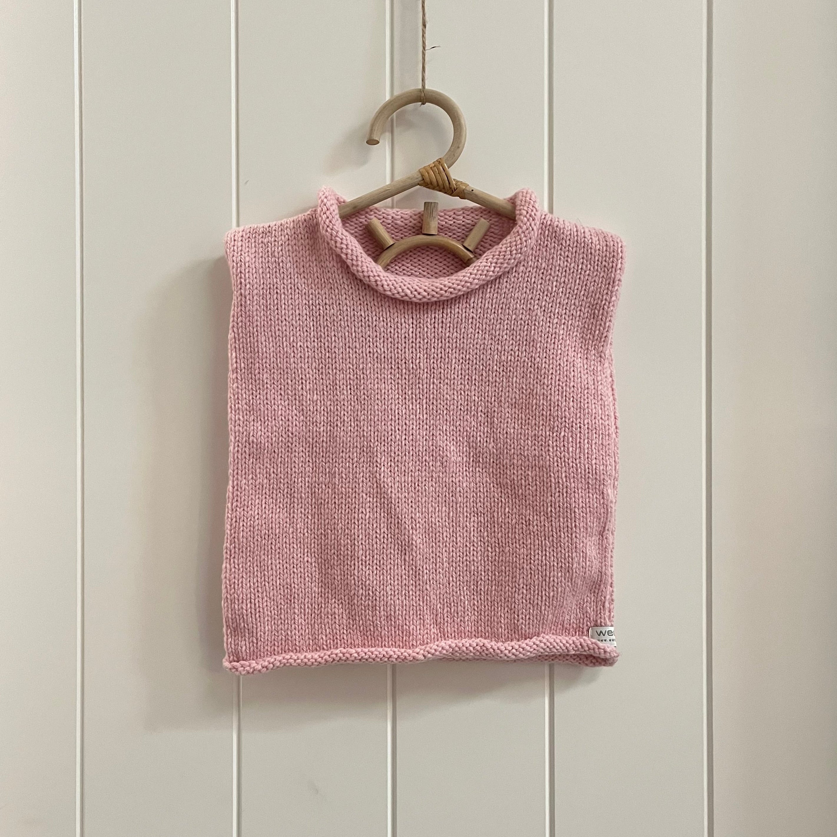 A pink knit sleeveless  vest hanging on a light wood hanger against a white paneled wall.