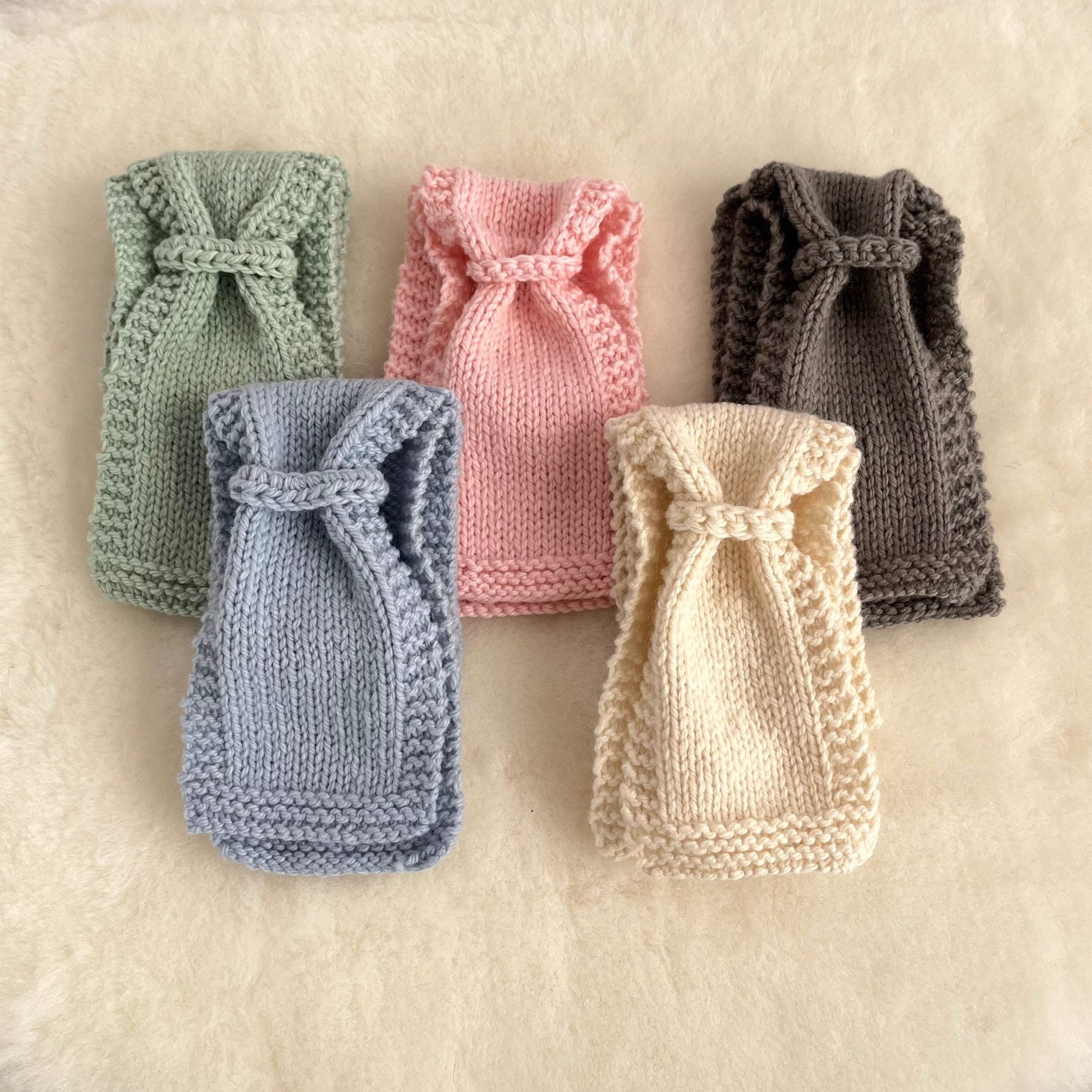 Five knitted baby scarves in pastel and neutral colors, arranged on a soft cream fabric background. The colors include sage green, dusty blue, pink, charcoal gray, and cream.