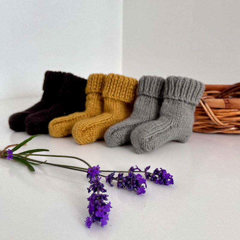 Three pairs of knitted baby socks in mustard, chocolate and mushroom with a wicker basket and lavender flowers on a white background.