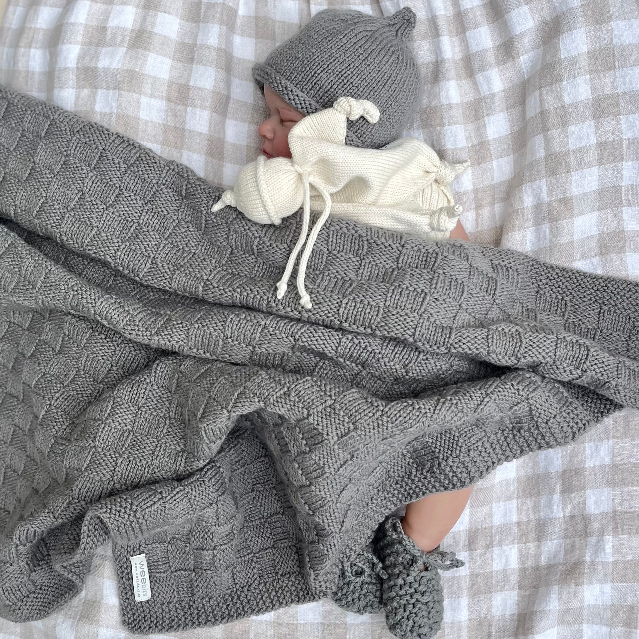 Baby wrapped in a mushroom knit travel rug with a matching beanie and booties on a checkered background.