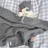 Baby wrapped in a mushroom knit travel rug with a matching beanie and booties on a checkered background.