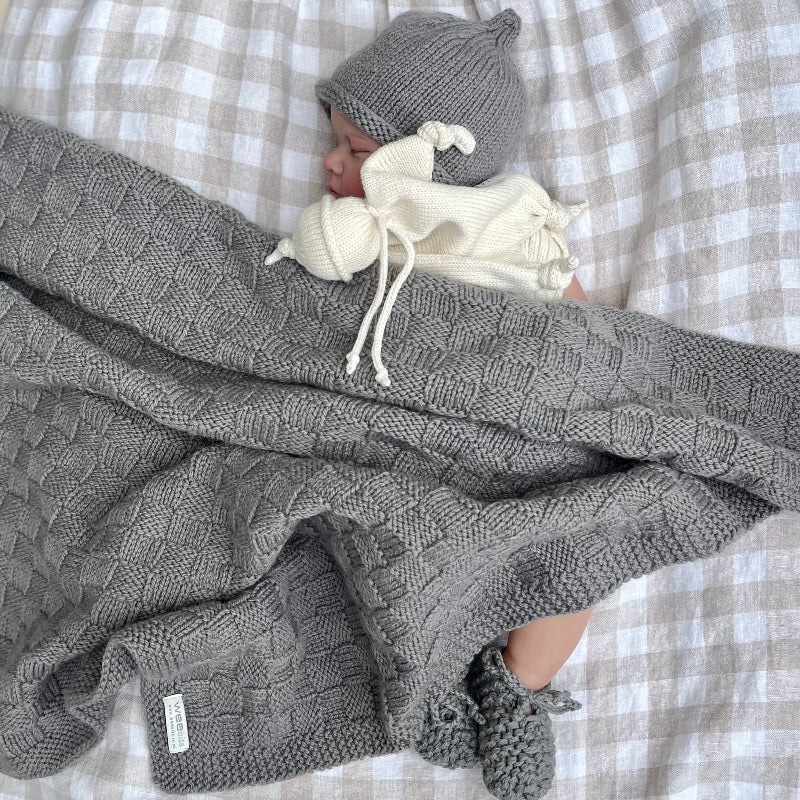 Baby wrapped in a mushroom knit travel rug with a matching beanie and booties on a checkered background.