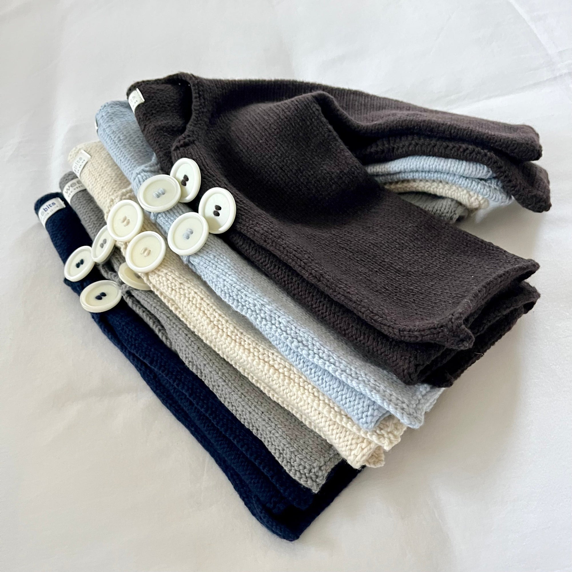 A stack of folded knit baby jackets in multiple colors, including dark brown, light blue, cream, gray, navy, and white, arranged on a white fabric background with decorative white buttons on top.