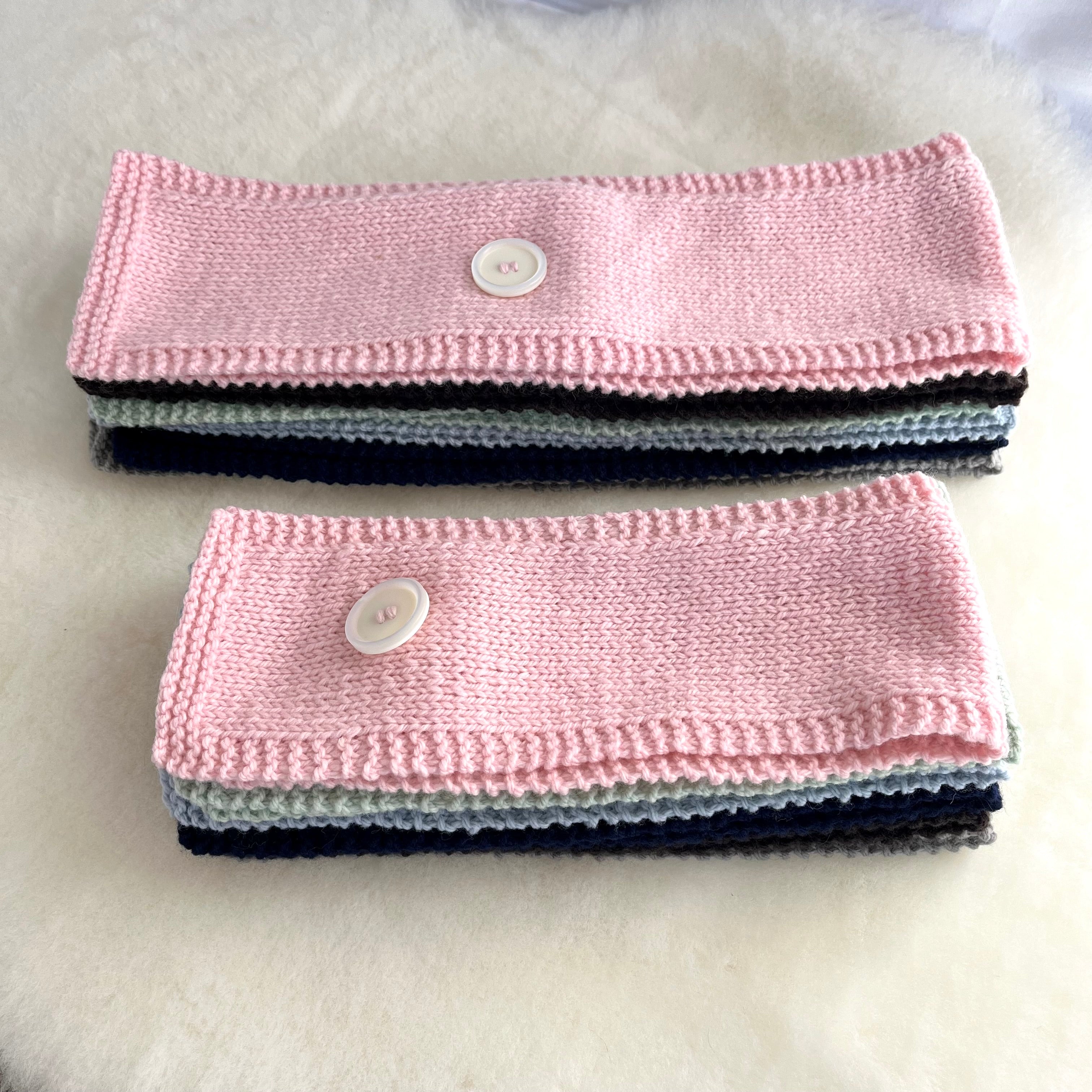 Two knitted scarf stacked on a soft white surface. Each scarf is light pink with a white button closure and shows a stack of other colors underneath, including black, light blue, and white.
