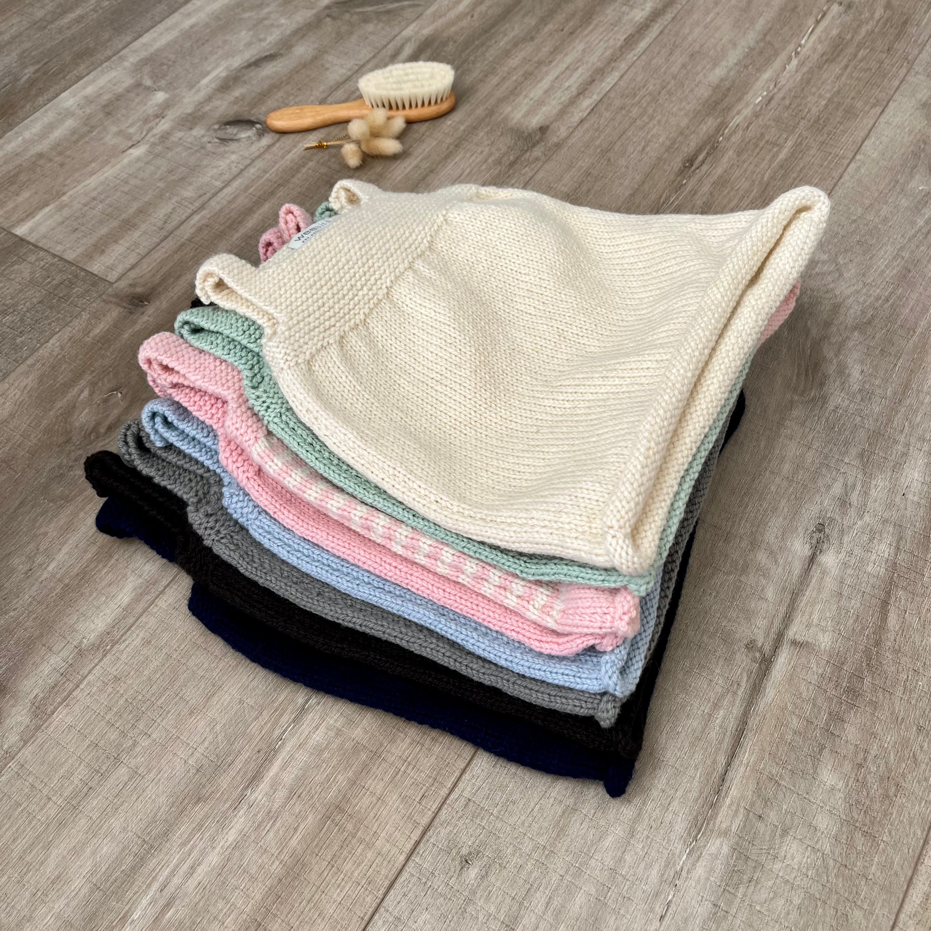A stack of folded knit baby dresses in multiple pastel and neutral colors, including cream, pink, mint, light blue, gray, and black, arranged on a gray wood floor with a small wooden brush nearby.