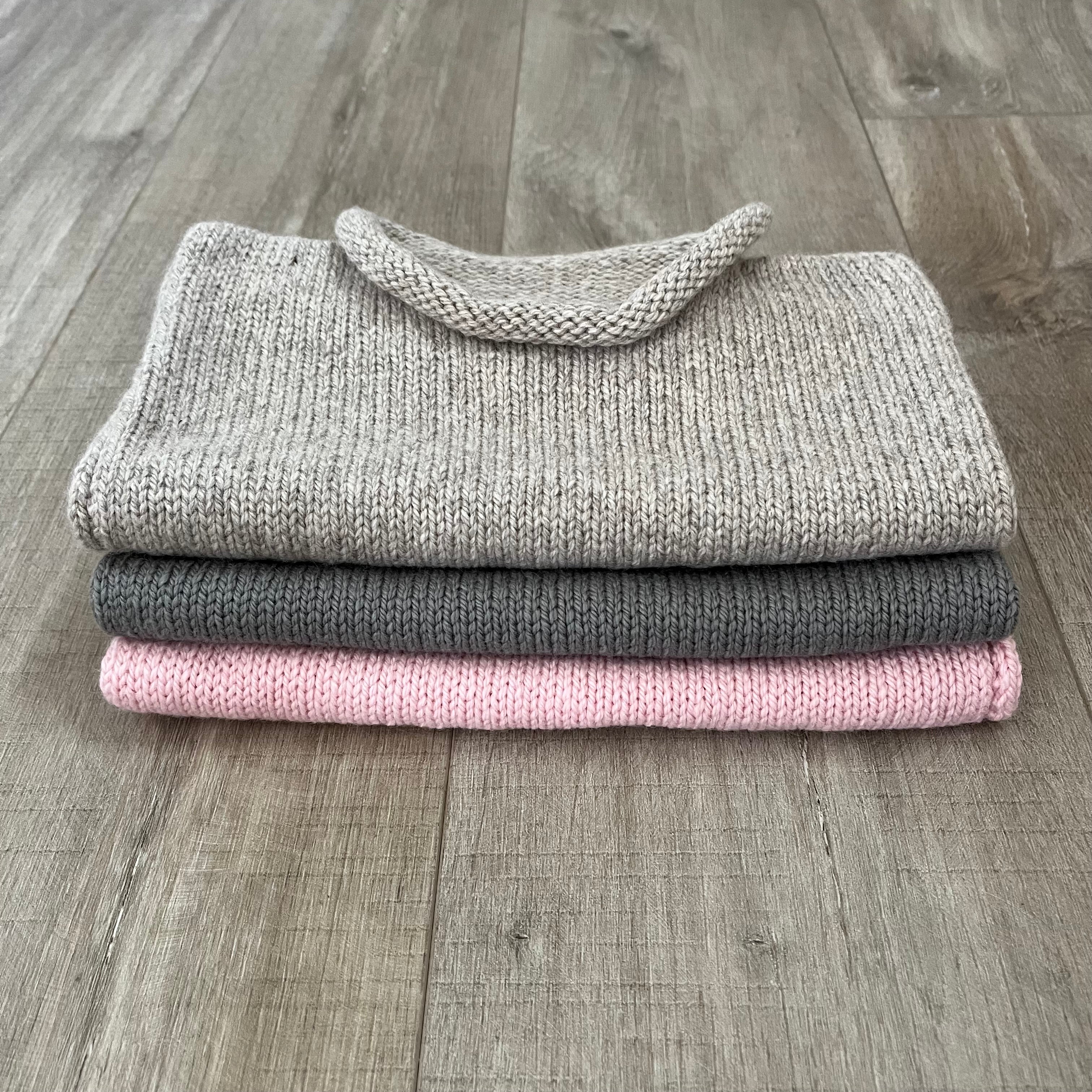 Three folded ribbed knit sweaters stacked on a gray wood floor, in light gray, dark gray, and soft pink.