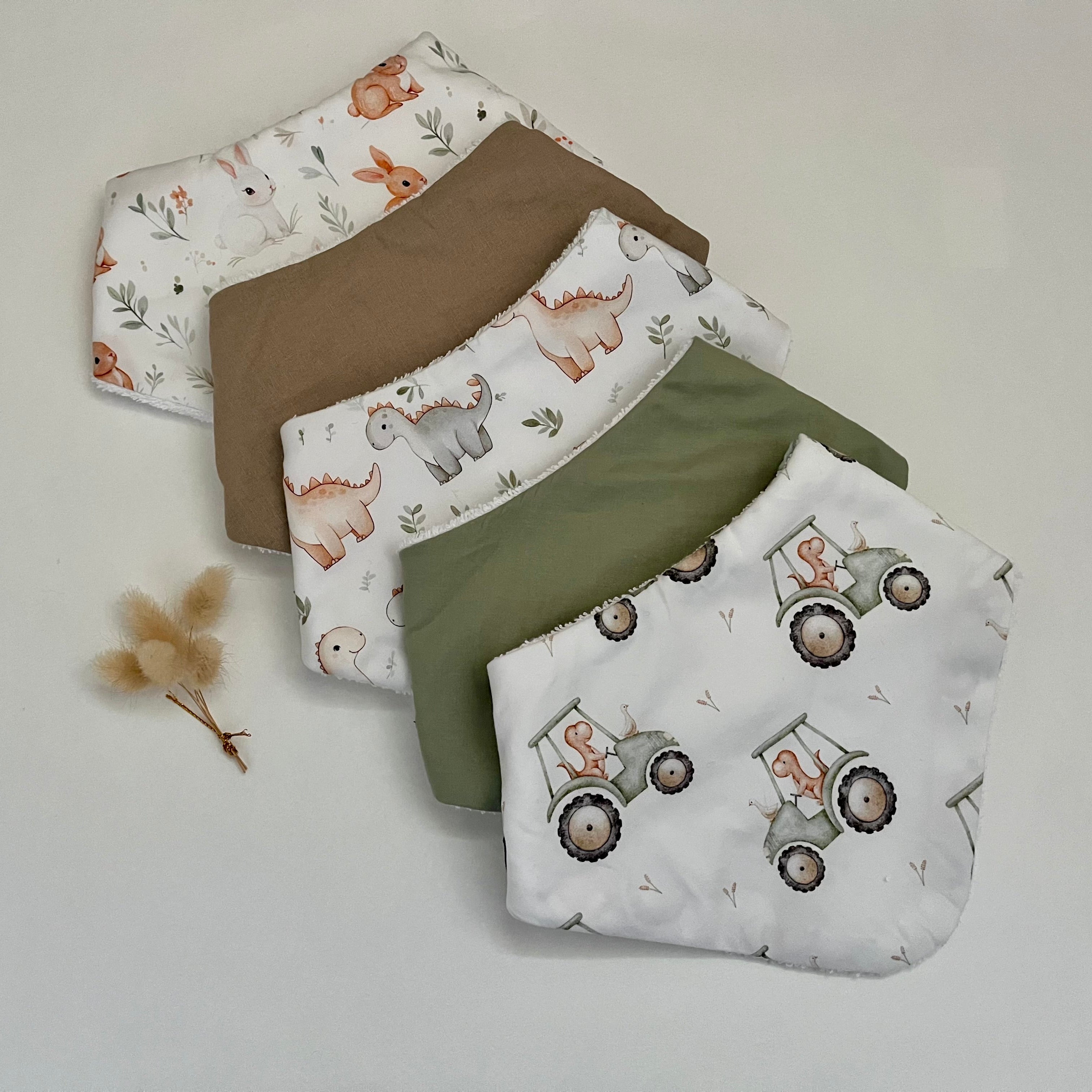 Five reusable cloth feeders or bib-style protectors arranged in a fan on a light neutral background. The fabrics include white with dinosaur prints, solid brown, white with tractor prints, solid olive green, and a white fabric with a small pattern.