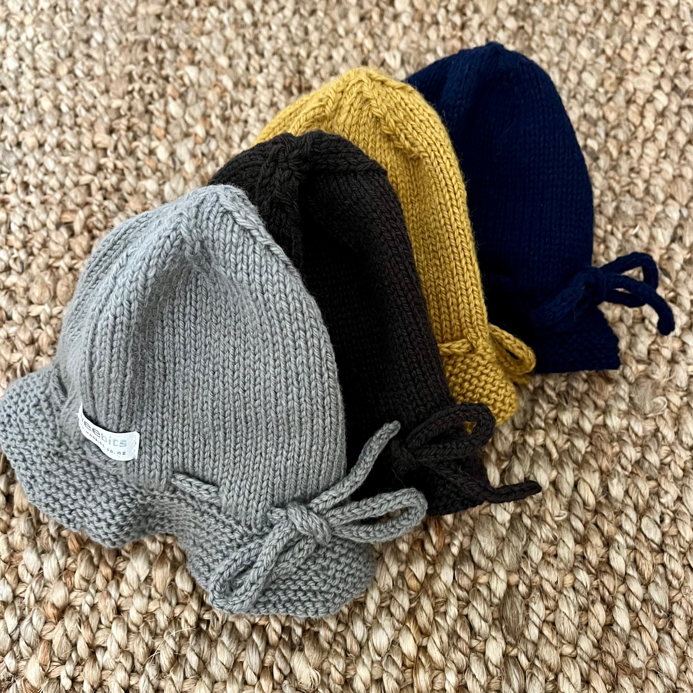 Four hand-knit baby hats in gray, brown, navy and mustard yellow, arranged diagonally on a textured beige woven rug. Each hat features a small bow on the front and a drawstring closure at the top.
