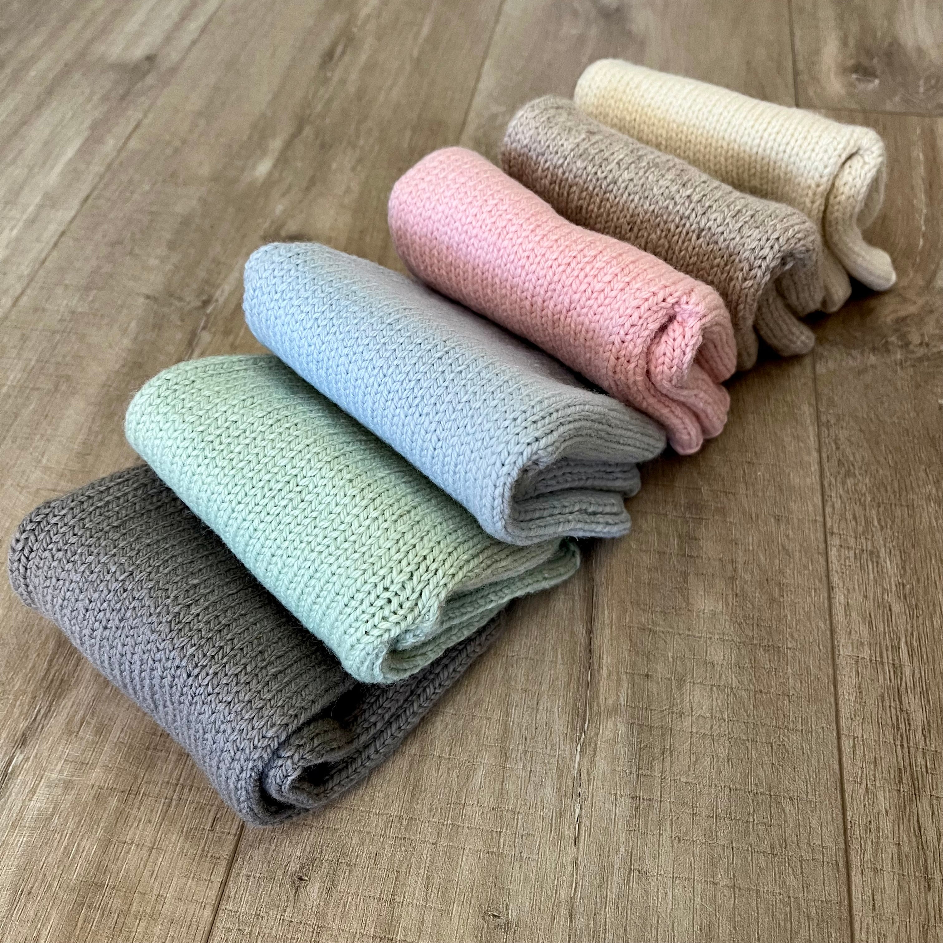 six rolled knit baby pants in muted pastel and neutral colors, arranged diagonally on a light brown wooden floor.