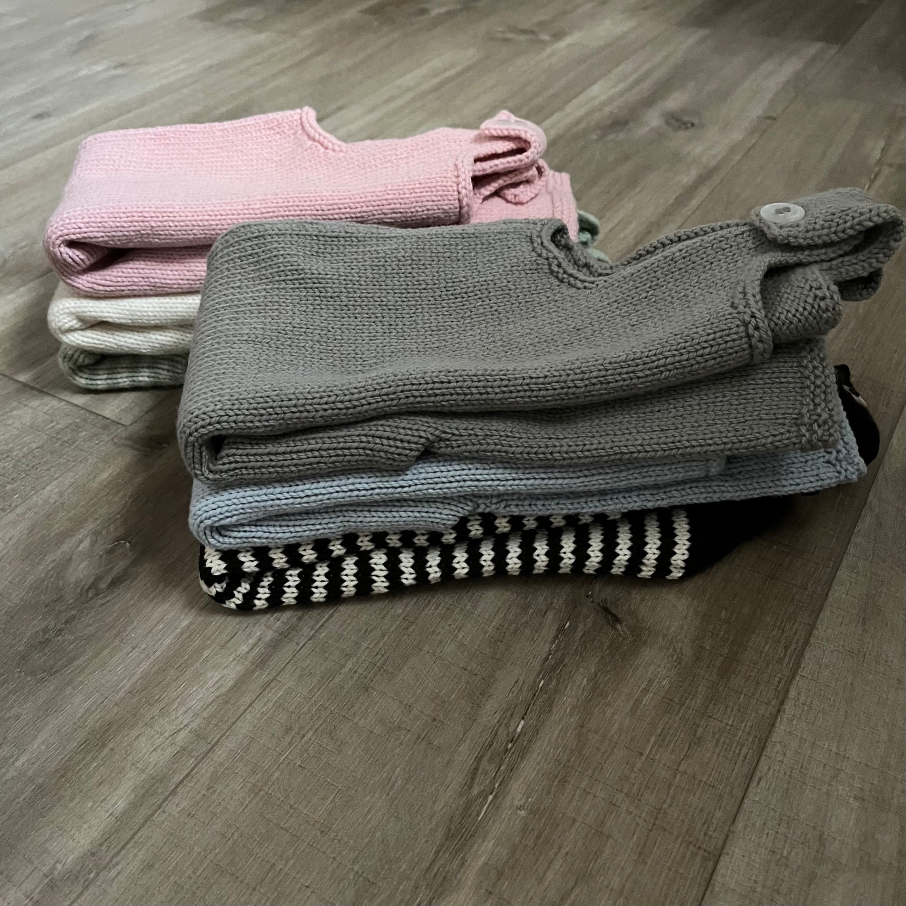 Three folded knit baby dungarees stacked on a wood floor. The front pair is a taupe-gray color with a small white button detail, the middle pair is light blue, and the back pair is black and white striped.