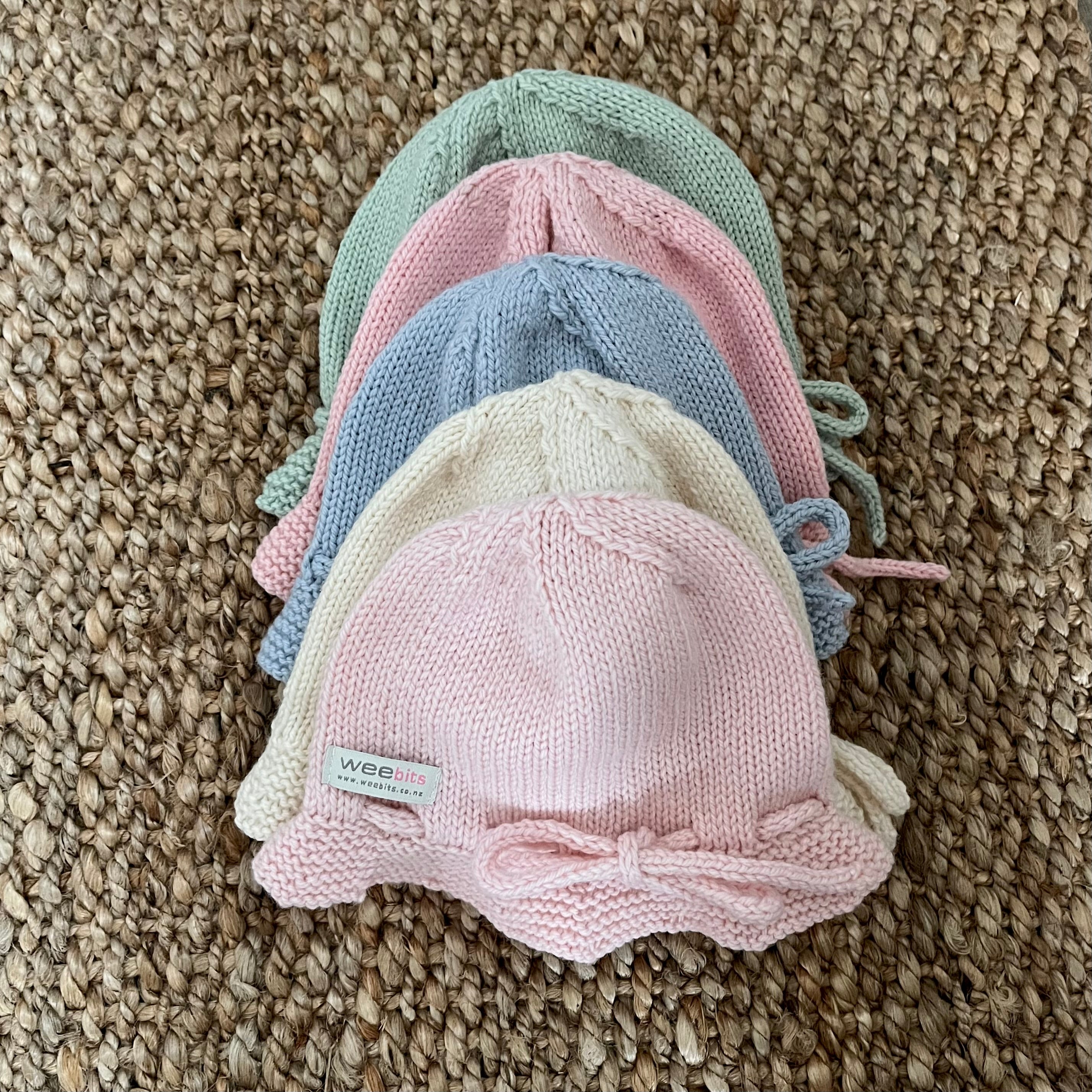 Four knitted baby hats in pastel shades of mint green, pink, light blue, and cream, stacked on a textured woven jute rug. The frontmost hat is light pink with a bow on the brim and a small white label that reads 'weebits'