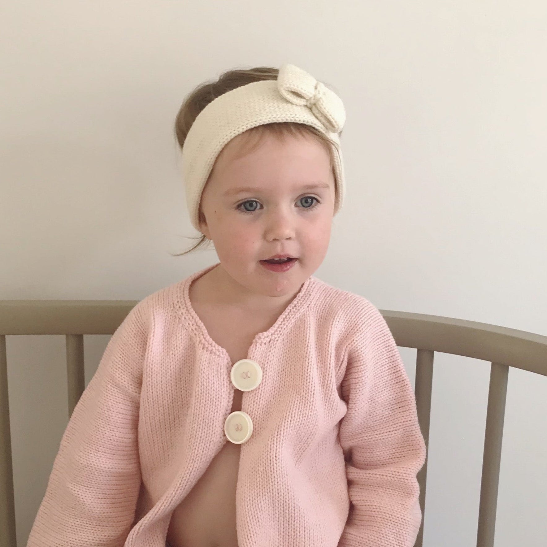 A toddler sitting in a round crib wearing a light pink knit cardigan with cream buttons, a cream bow headband