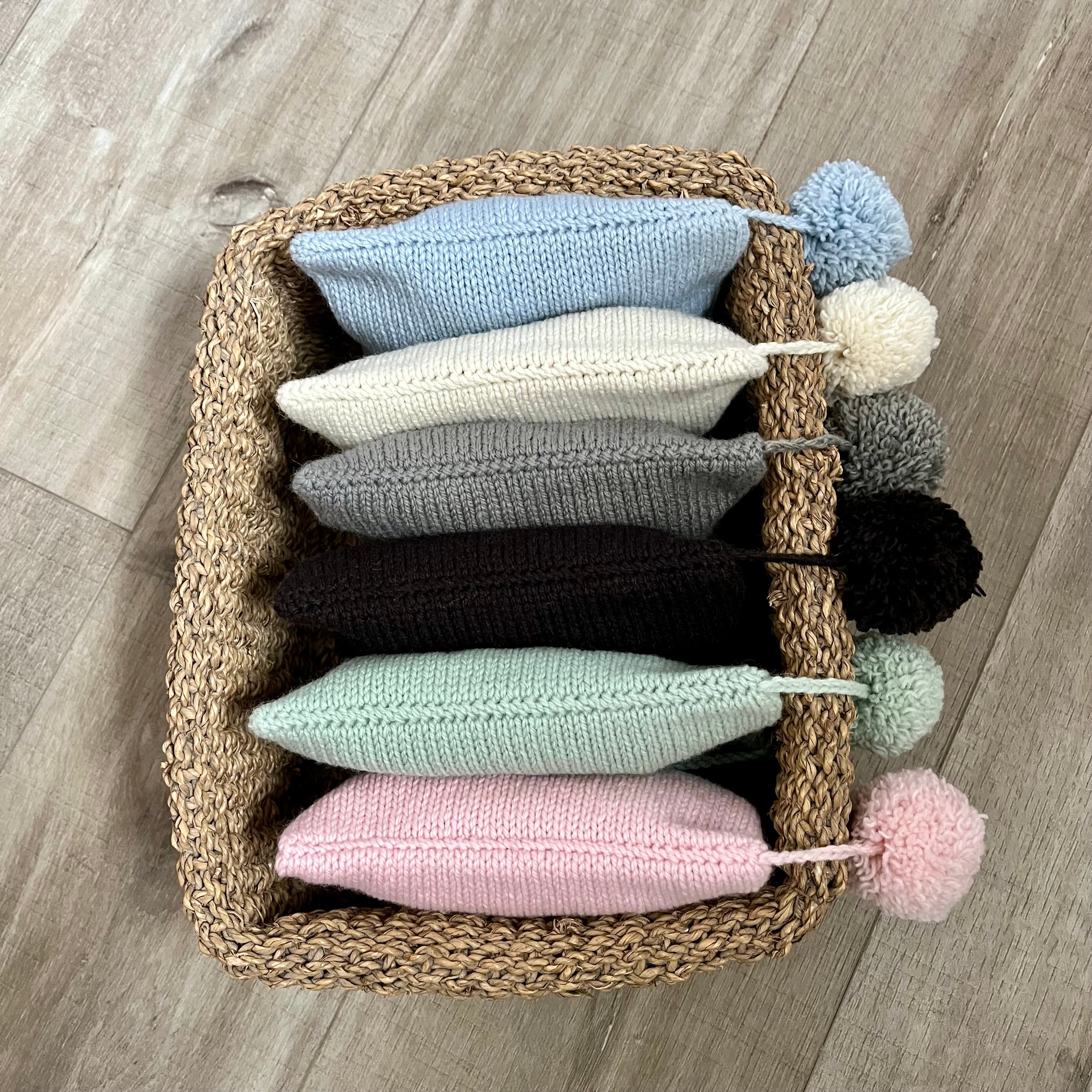 Six knitted pompom hats in pastel and neutral shades are arranged inside a woven straw basket on a light wood floor. Each hat has a pom-pom attached to the side.