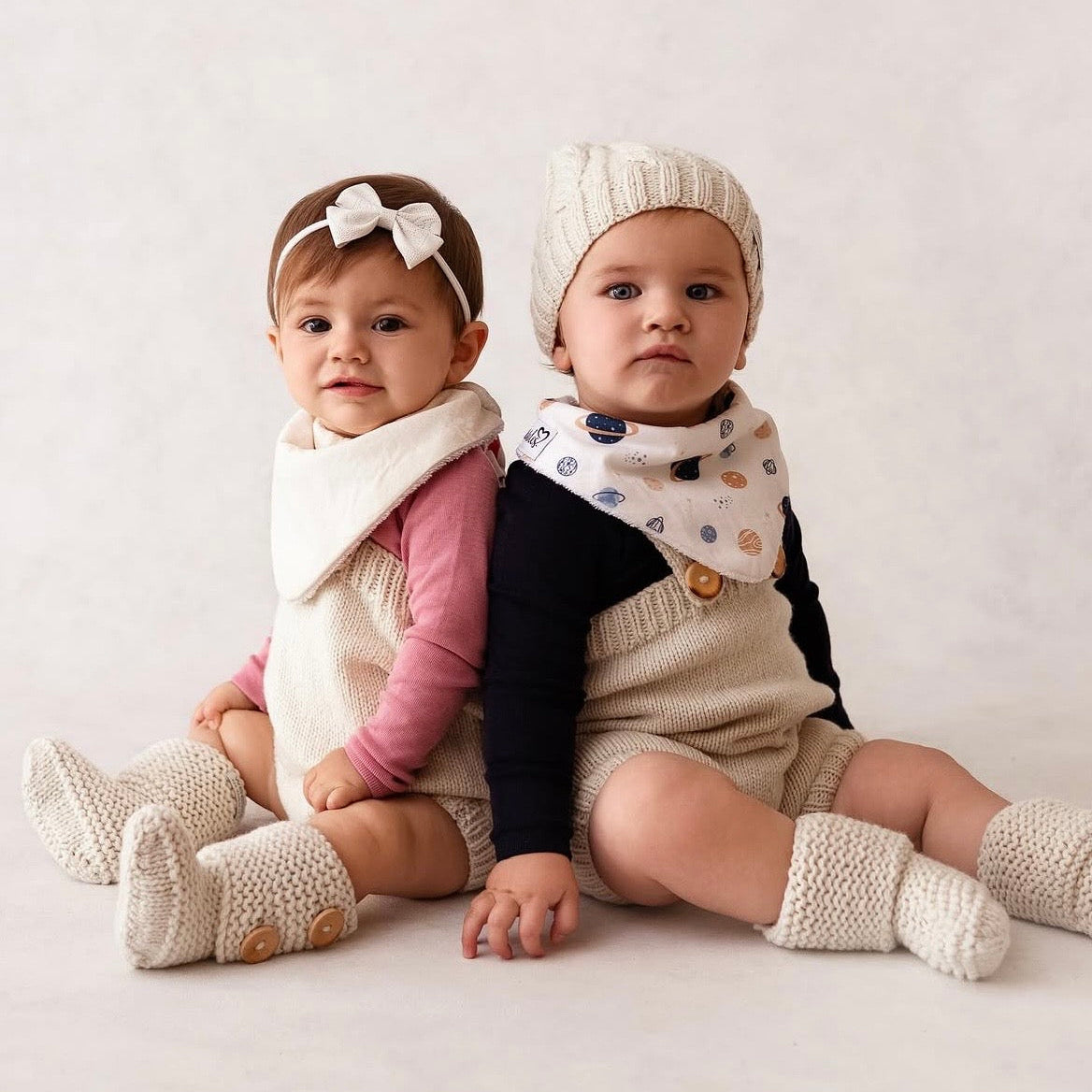 Two babies sitting back-to-back wearing matching knit baby clothing sets, including cream bibs, booties, and hats, against a soft off-white studio background.