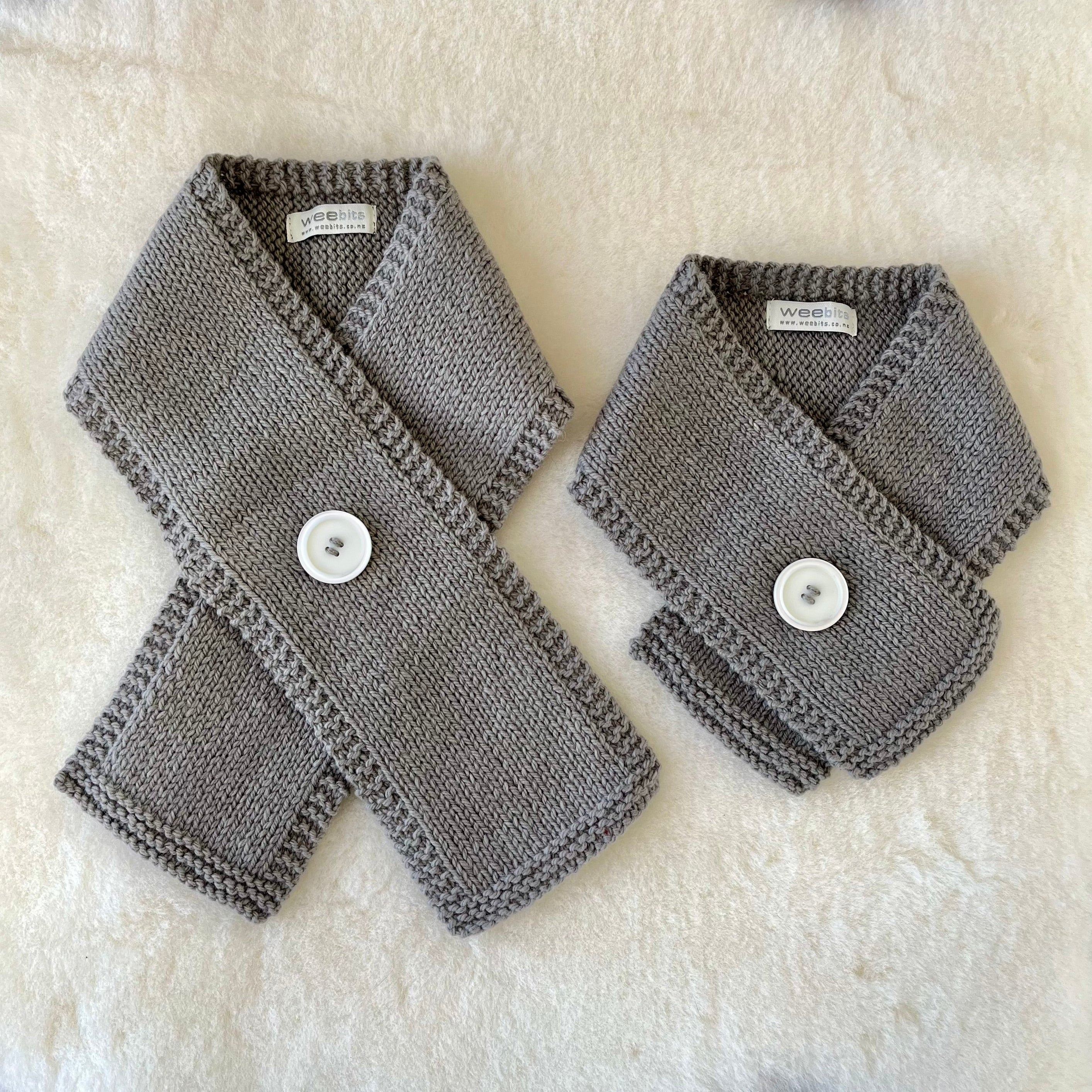 Two gray knitted button scarves with white buttons, displayed on a white faux fur surface.
