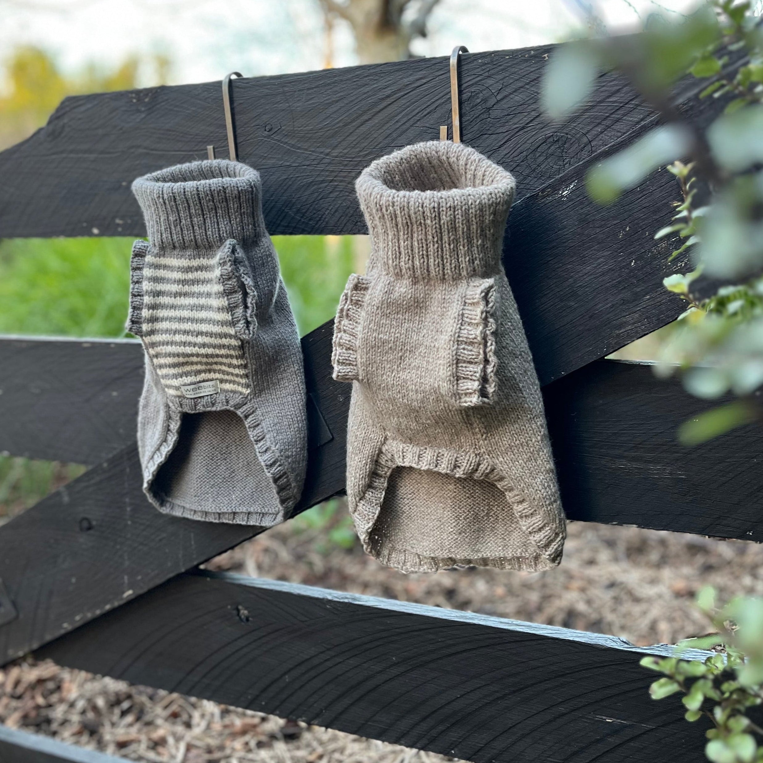 Two knit pet dog jumpers in natural beige and taupe hanging from a black wooden fence outdoors.